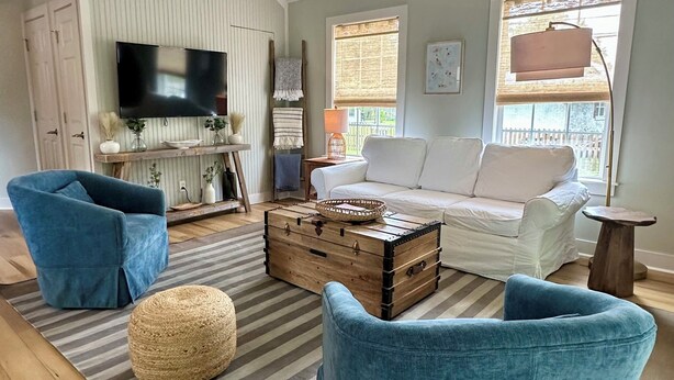 Un salon comprenant un téléviseur, un canapé, deux fauteuils bleus, une commode en bois et un tapis à rayures.