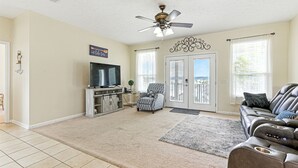 A living room with a television, a ceiling fan, a sofa, and a large window. - Compass Lake - Cypress Sunsets! (Alford)