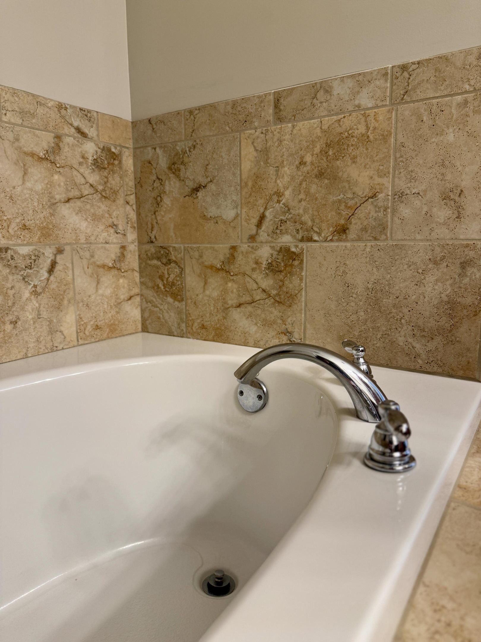 A bathroom with a white bathtub, a chrome faucet, and beige marble tiles on the walls.