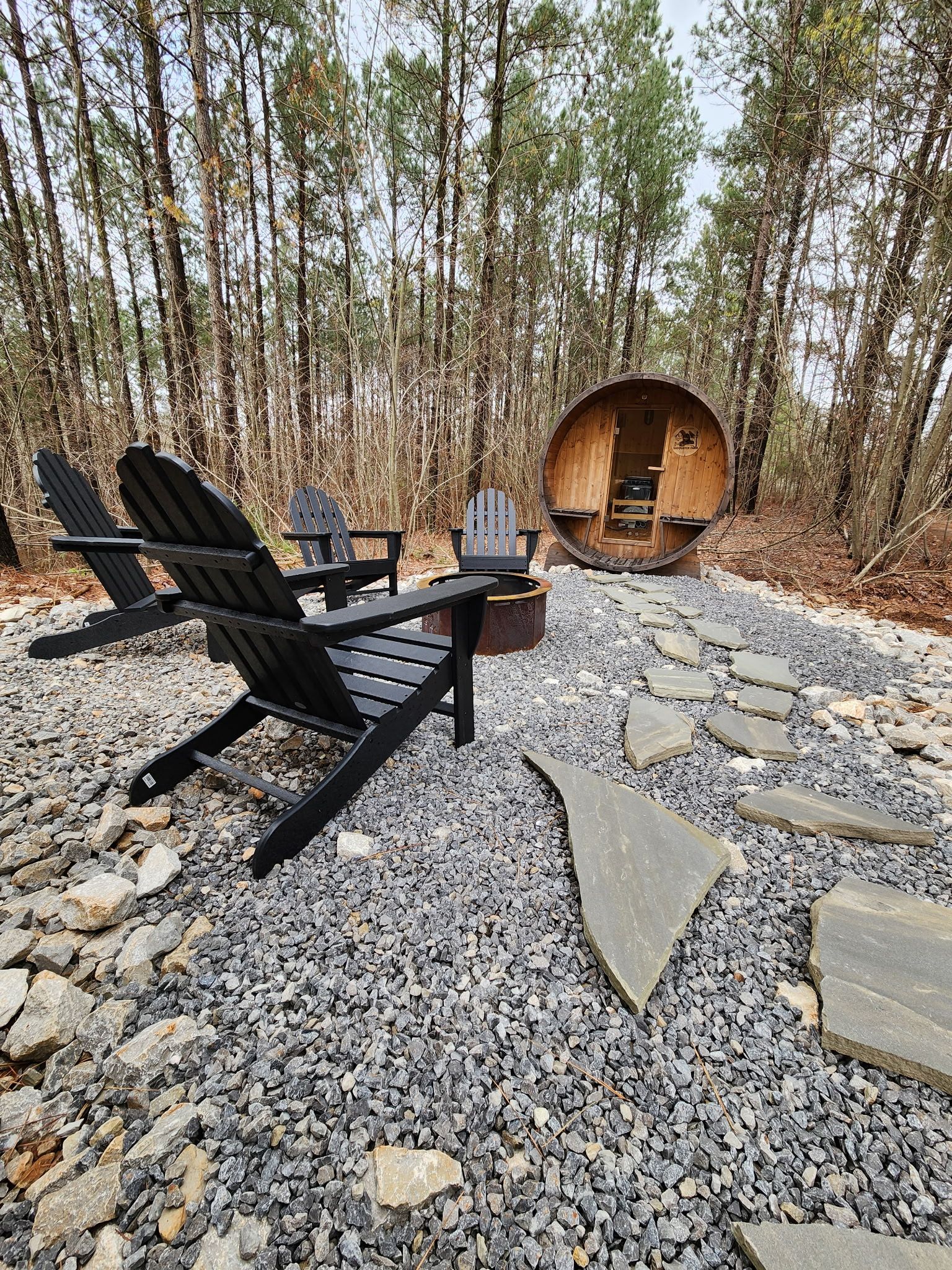 A rustic outdoor seating area with Adirondack chairs and a fire pit, surrounded by a forest.