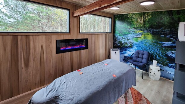 Une pièce aux murs en bois, un lit recouvert d’une courtepointe bleue, une table de massage, une cheminée fixée au mur et une grande fenêtre donnant sur la forêt.