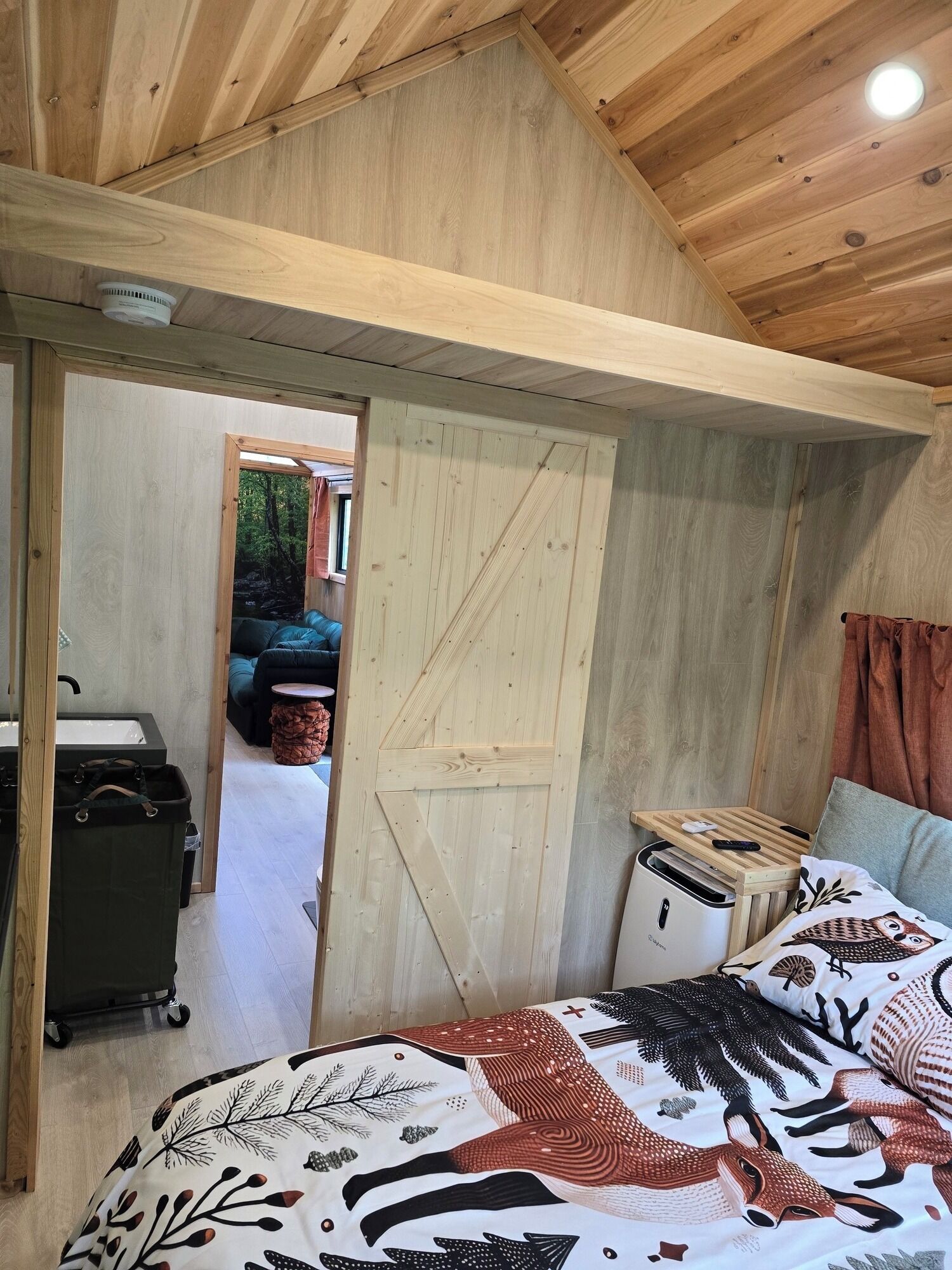 A bedroom with a wooden ceiling, a bed with a patterned blanket, a small table with a remote, and a door leading to an outdoor area.