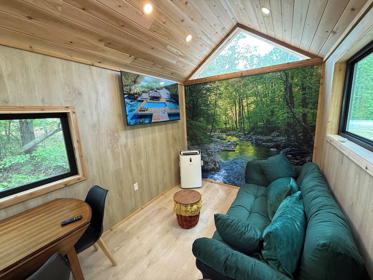 Interior acolhedor de uma cabana, com teto de madeira, sofá verde, mesa pequena e TV exibindo uma cena tropical.