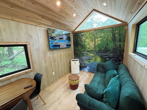 Un acogedor interior de cabaña con techo de madera, un sofá verde, una mesita y un televisor que muestra una escena tropical.