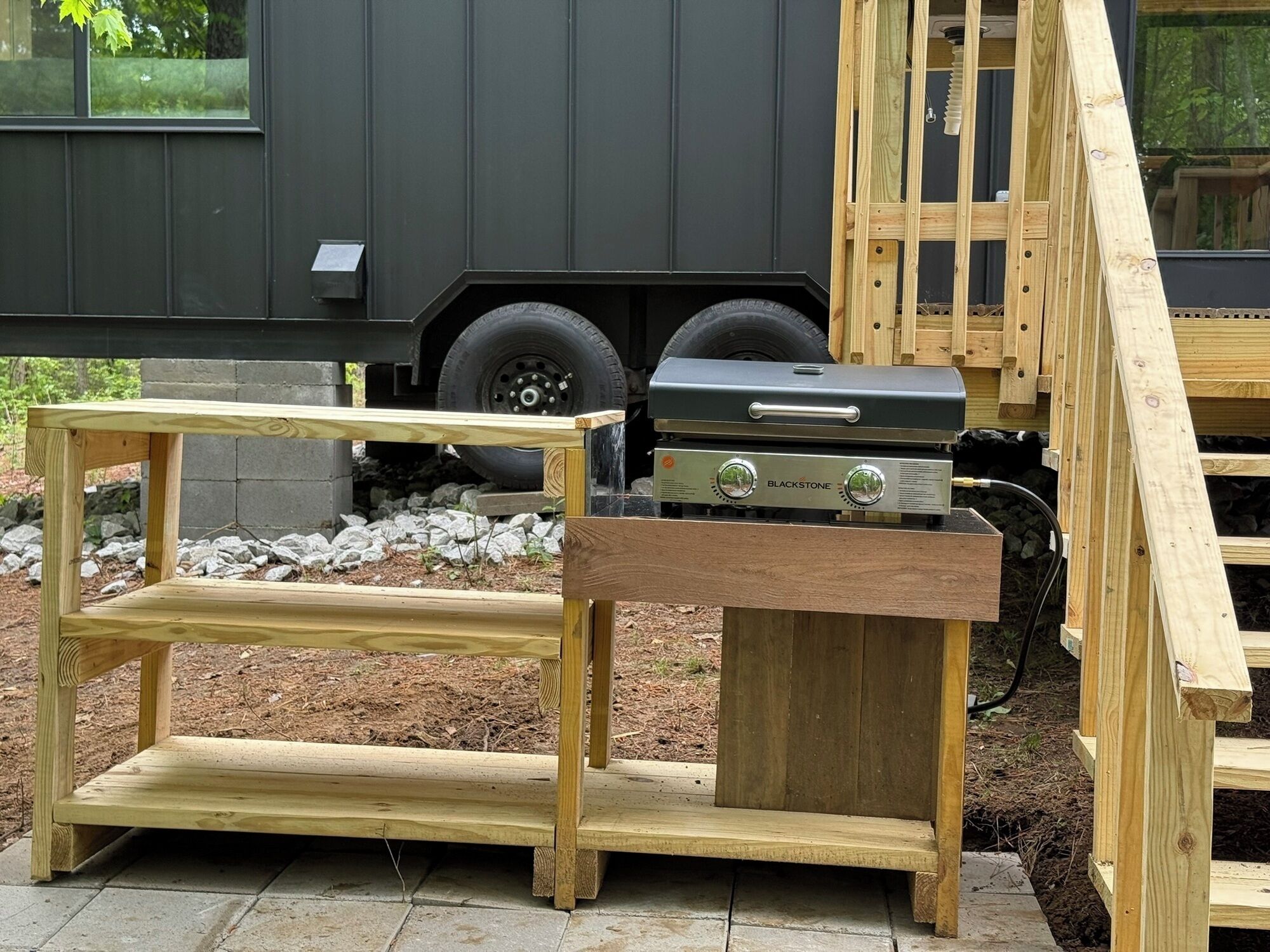 Una estructura de madera al aire libre con una parrilla y una mesa, frente a un edificio con un vehículo al fondo.