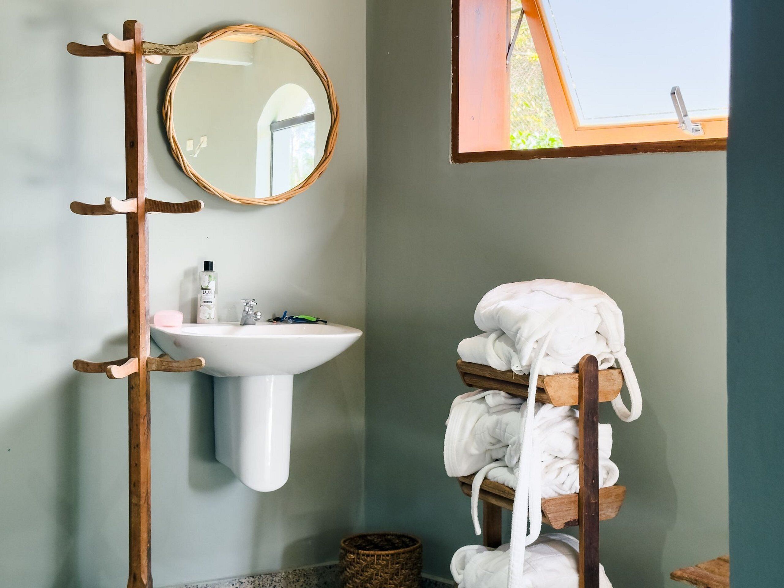 A bathroom with a wooden towel rack holding white towels, a round mirror, and a white sink with toiletries.
