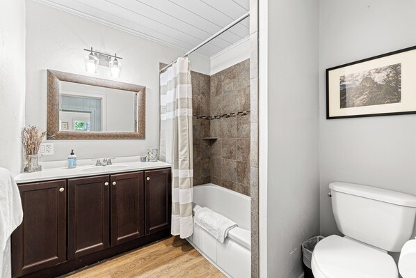 A bathroom with a double sink, dark wooden cabinets, a white toilet, and a shower with a stone wall. - Farmhouse on Main - historic home in the heart of Riggins (Riggins)
