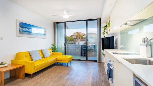A modern apartment with a yellow sofa, a dining table, a kitchen with a TV, and a sliding glass door leading to a balcony. - Scarborough Pearl Apartment (Scarborough)