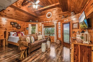 A log cabin interior with a wooden ceiling, a bed, a sofa, a TV, and a large window. - Lookout Point - Firepit, Pickle Ball, Balcony, Pet (Sevierville)
