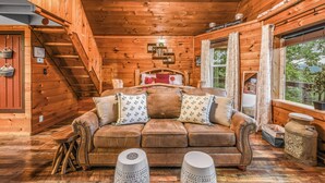 A wooden cabin interior with a sofa, pillows, a small table, and a window with curtains. - Lookout Point - Firepit, Pickle Ball, Balcony, Pet (Sevierville)