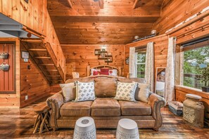 A wooden cabin interior with a sofa, pillows, a small table, and a window with curtains. - Lookout Point - Firepit, Pickle Ball, Balcony, Pet (Sevierville)