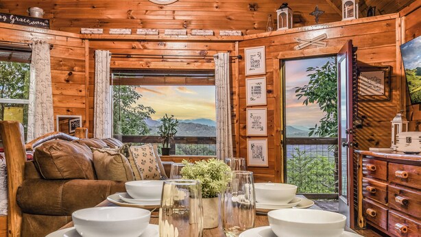A wooden cabin interior with a dining table set, a brown leather sofa, and a view of the outdoors through a window. - Lookout Point - Firepit, Pickle Ball, Balcony, Pet (Sevierville)