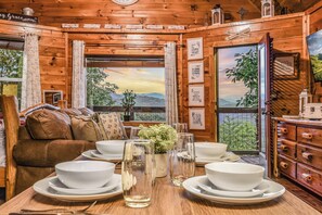 A wooden cabin interior with a dining table set, a brown leather sofa, and a view of the outdoors through a window. - Lookout Point - Firepit, Pickle Ball, Balcony, Pet (Sevierville)