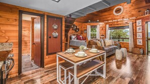 Interior de una cabaña de madera con una mesa de comedor, sofá y una escalera que sube a un altillo.
