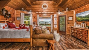 Interior de una cabaña de madera con cama, sofá, cómoda de madera y un amplio ventanal con vistas panorámicas.