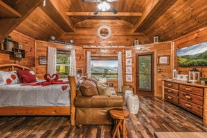 A wooden cabin interior with a bed, sofa, wooden dresser, and a large window offering a scenic view. - Lookout Point - Firepit, Pickle Ball, Balcony, Pet (Sevierville)