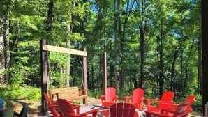 Une terrasse en bois, agrémentée de chaises Adirondack rouges et d’un banc en bois, le tout entouré d’arbres.