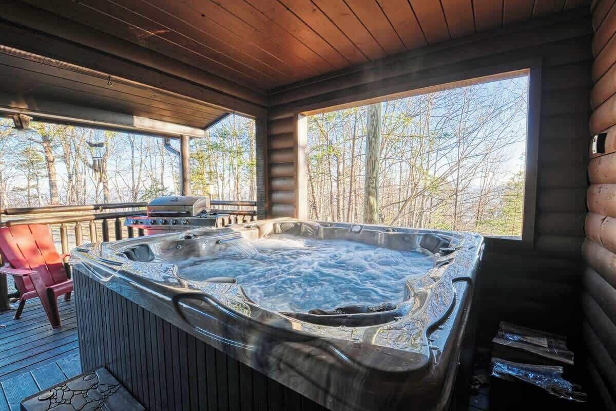A hot tub in a wooden cabin with a deck, a grill, and a view of trees.
