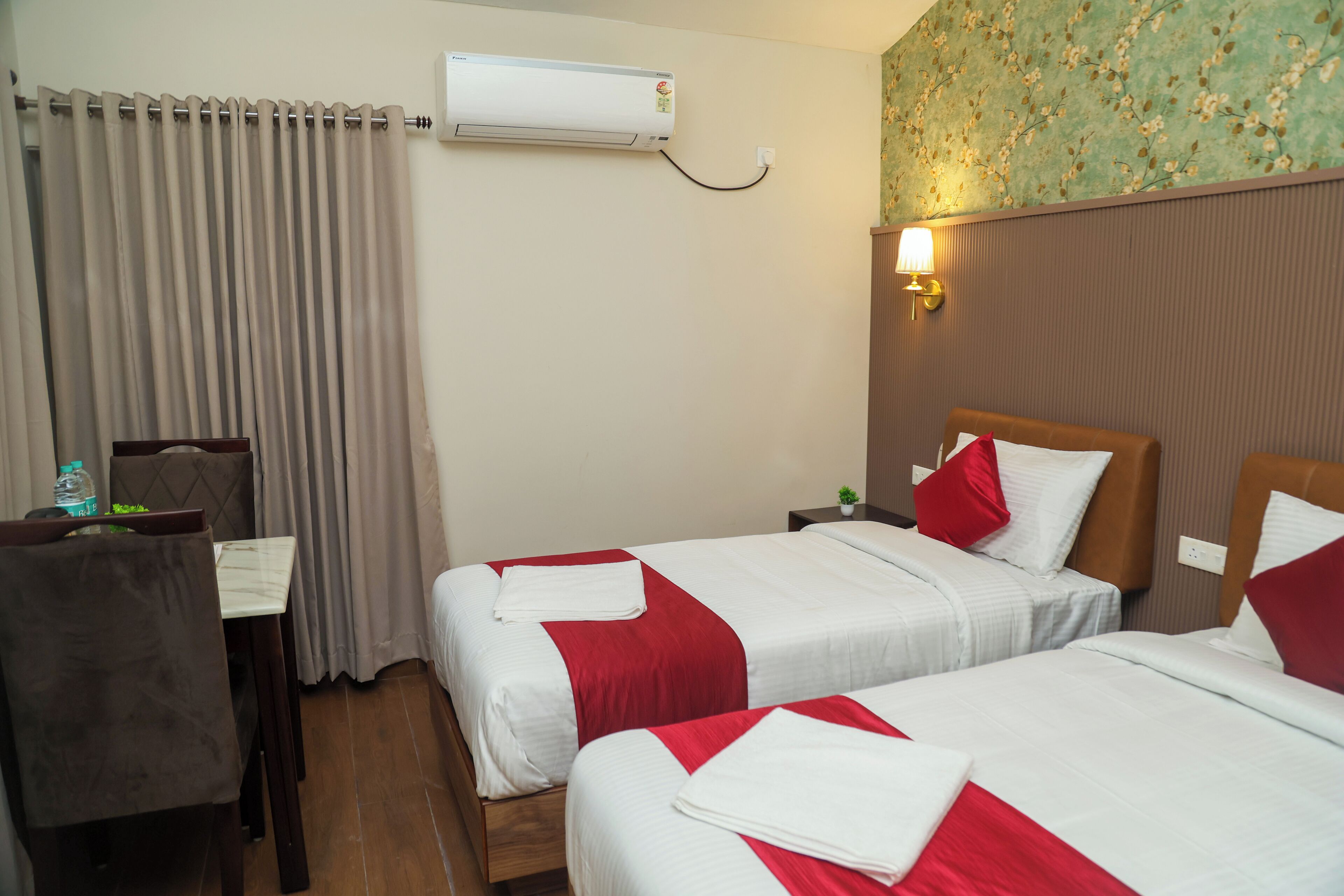 A hotel room with two beds, a desk, a chair, an air conditioner, and a wall-mounted lamp.