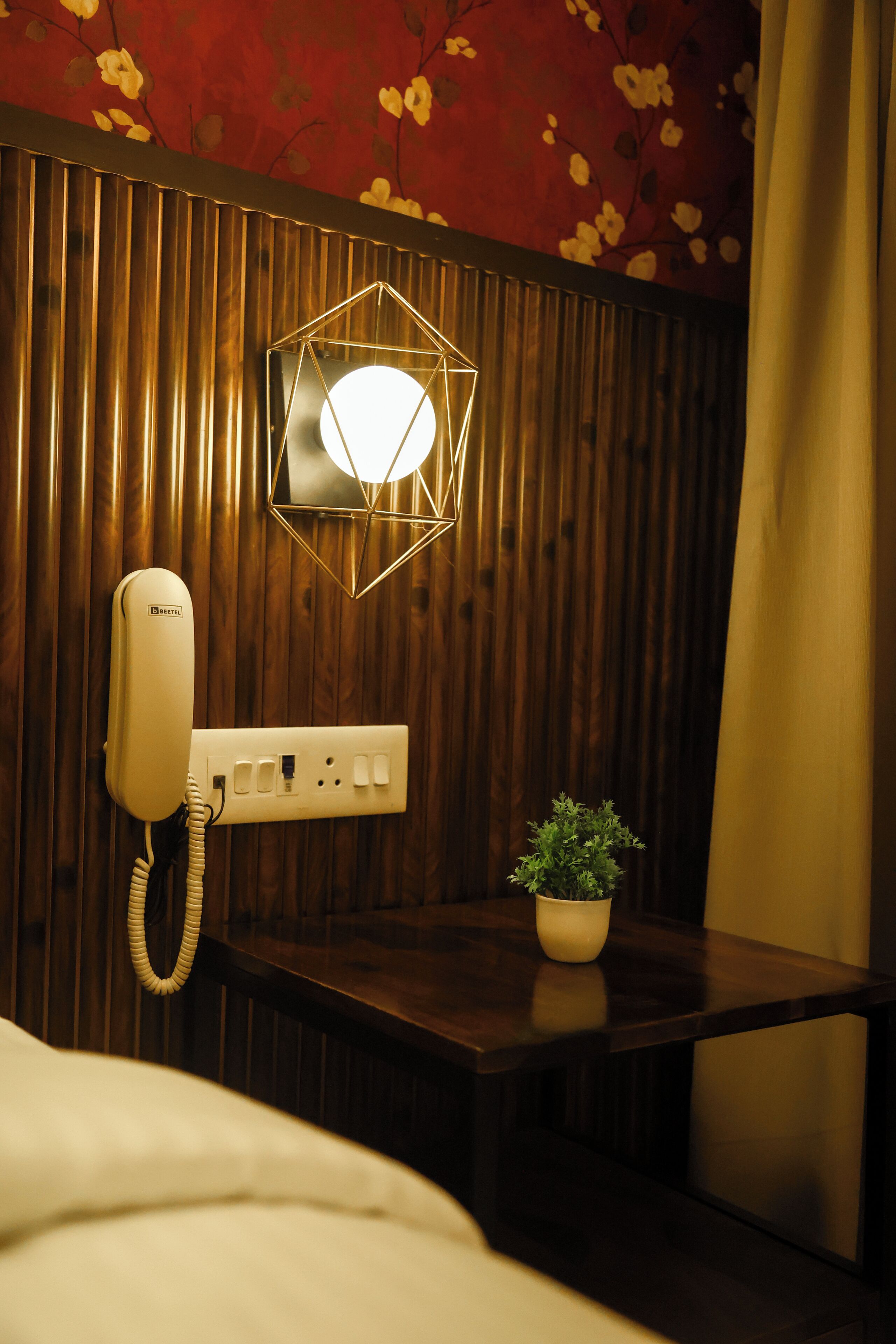 A hotel room with a wooden panel wall, a white telephone, a wall-mounted light fixture, and a small potted plant on a wooden nightstand.