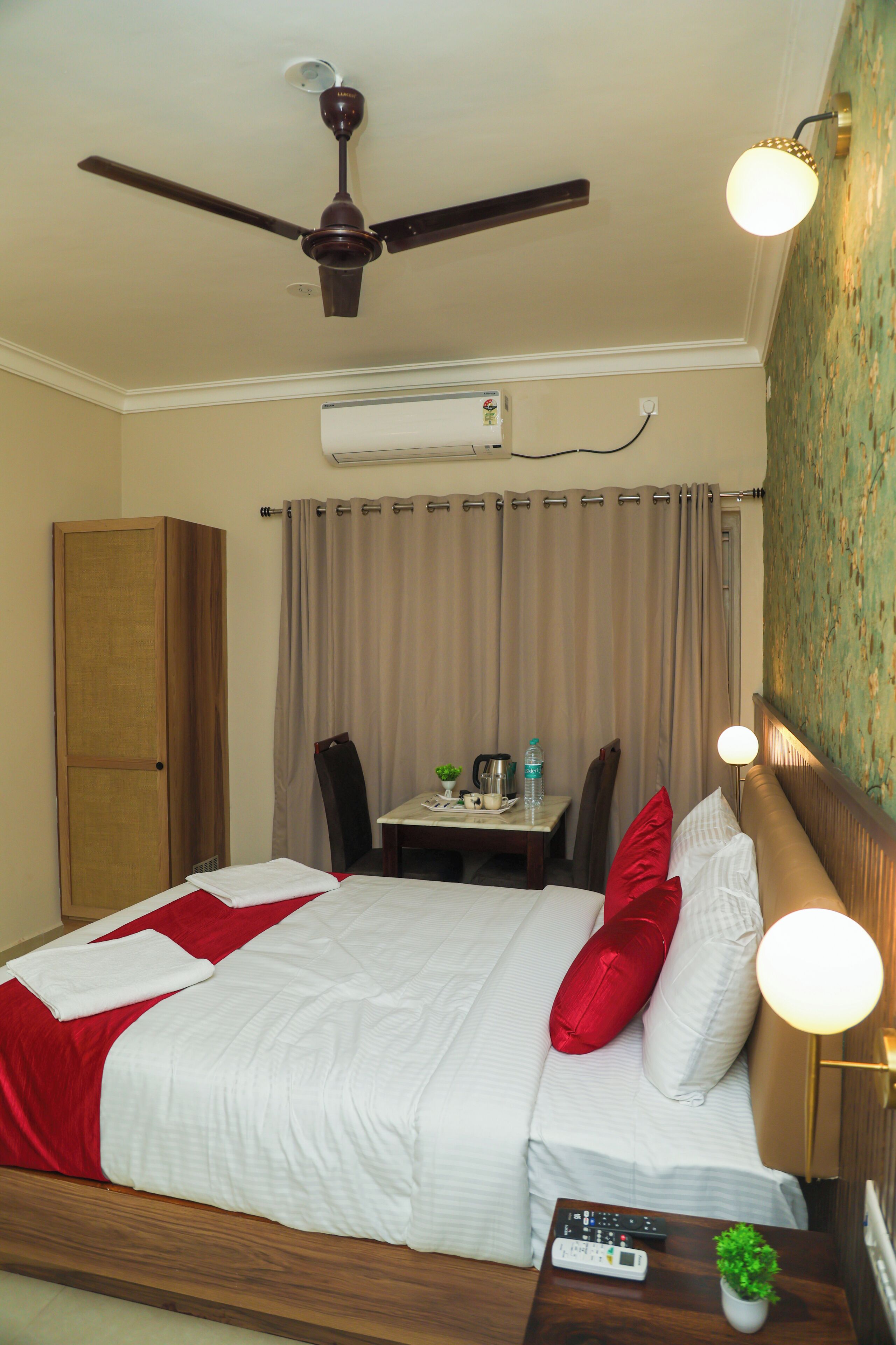 A hotel room with a bed, a desk, a chair, a ceiling fan, an air conditioner, and a nightstand with a remote control and a small plant.
