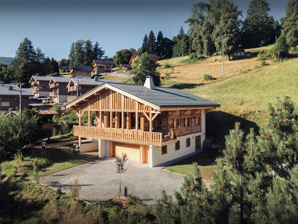 Una cabaña de madera con balcón, rodeada de árboles y otras casas en una zona montañosa.