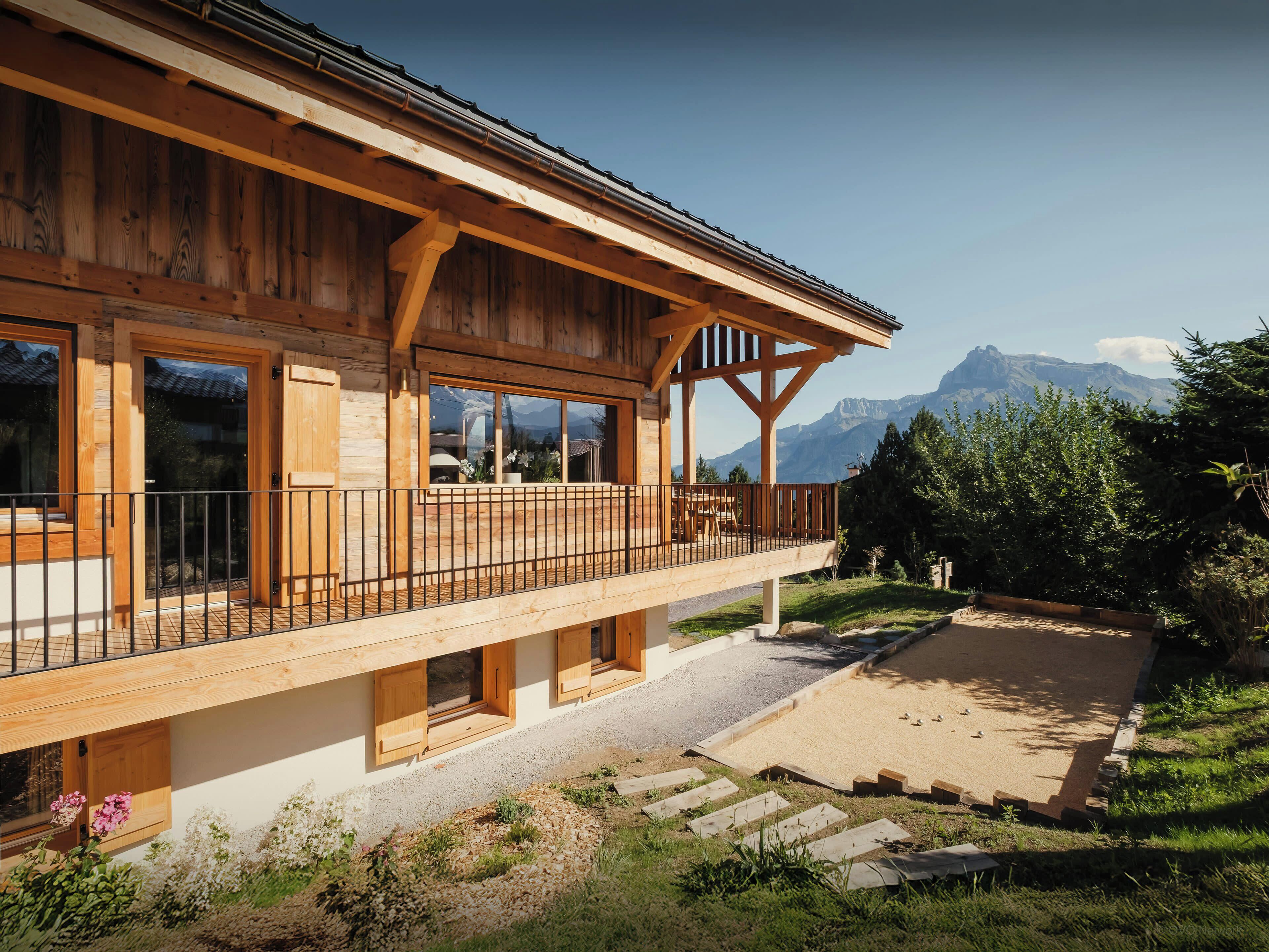 A wooden chalet with a balcony, a gravel driveway, and a mountainous backdrop.