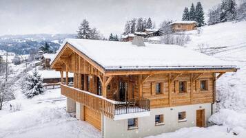 A wooden chalet with a balcony, surrounded by snow-covered trees and hills.