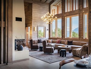 Un salón amplio con un sofá grande, sillones, una mesa de centro y una mesa de comedor preparada para dos. Hay una araña colgando del techo y ventanas grandes que ofrecen vistas al exterior.