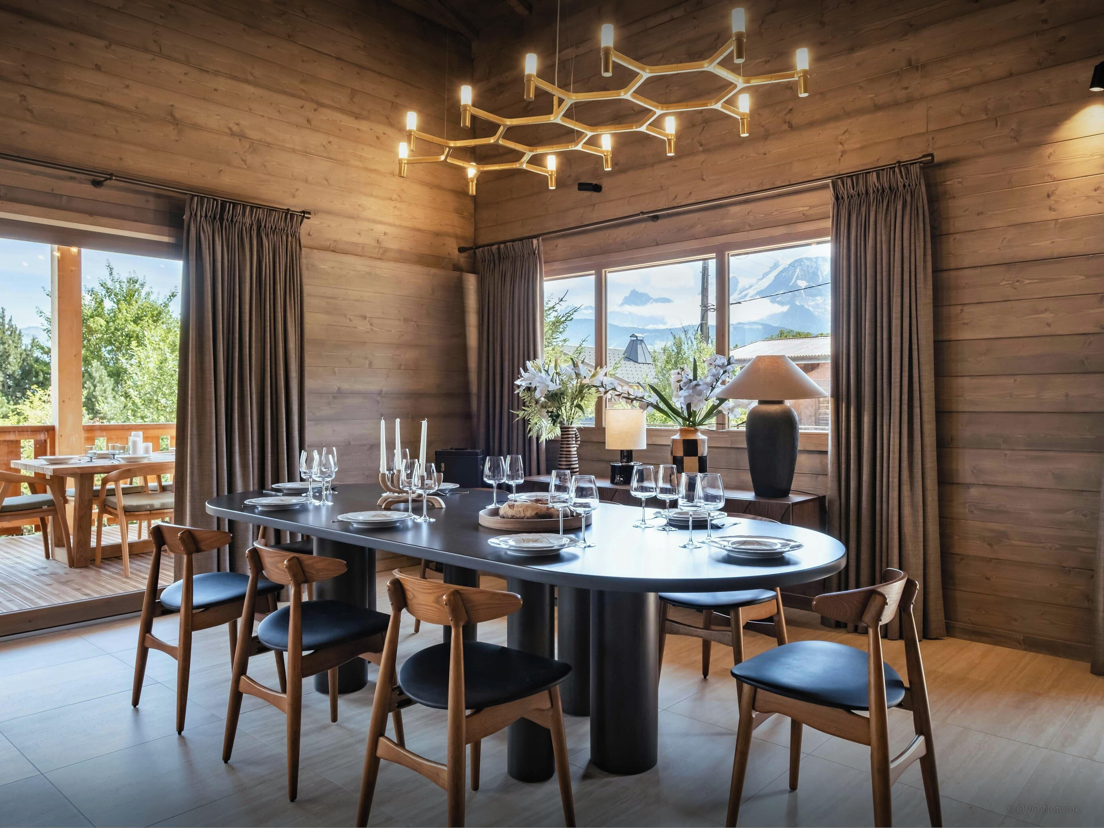 Un coin repas avec une table ronde, des chaises, des bougies et une vue sur les montagnes, visible à travers de grandes fenêtres.
