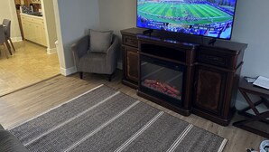 Una sala de estar con una televisión de pantalla plana, un sillón cómodo, una chimenea y una cocina visible a través de un arco.