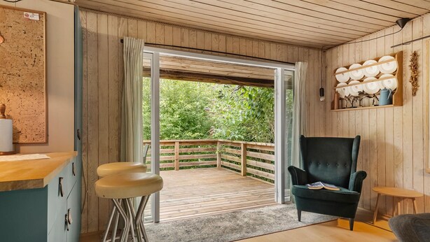 A room with a wooden ceiling, a wooden-framed door leading to a balcony, a dark green armchair, a small wooden table, and a bar stool.