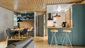 A modern kitchen with a wooden ceiling, a dining area with a grey sofa, and a bar area with stools.