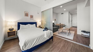 A modern hotel room with a large bed, a desk, and a sliding glass door leading to a living area.