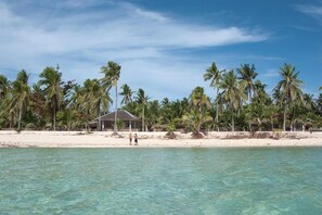 Een strand met helder turquoise water, palmbomen en een klein gebouw in de verte.
