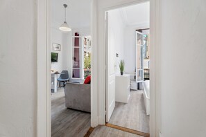 A hallway with a grey sofa, a dining area with a table and chairs, and a view of a balcony with plants. - Appartement Perle - Welkeys (Nice)