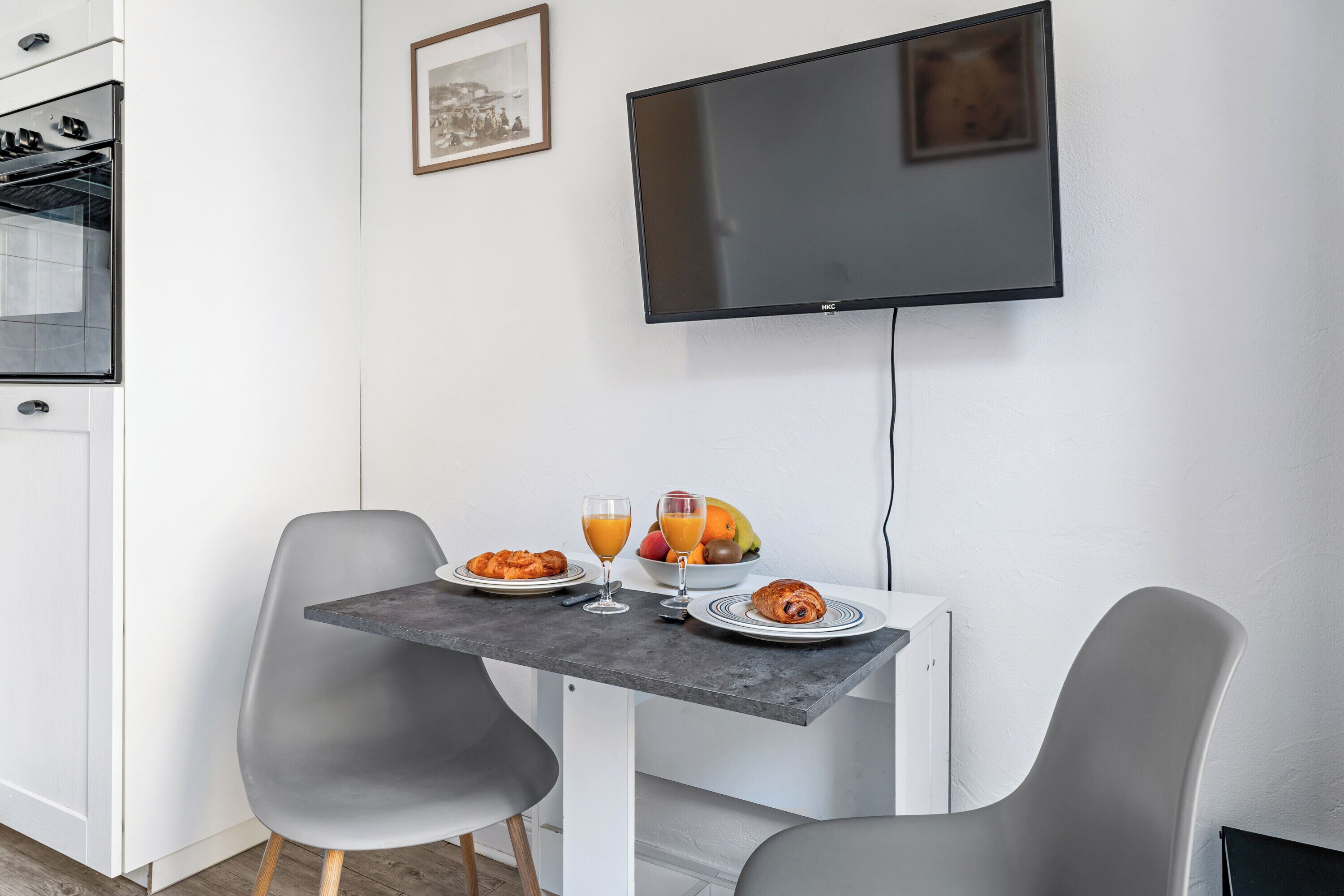 A modern kitchen with a flat-screen TV, a dining table with two chairs, and a breakfast setting with croissants and fruit.