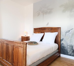A bedroom with a wooden bed, white bedding, a nightstand with a lamp, and a wall mural of tropical plants.