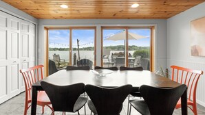 A dining area with a large table and chairs, a wooden ceiling, and large windows with a view of the outdoors. - White Lake MI Lakeside Home, Hot Tub, Pet Friendly (White Lake Township)