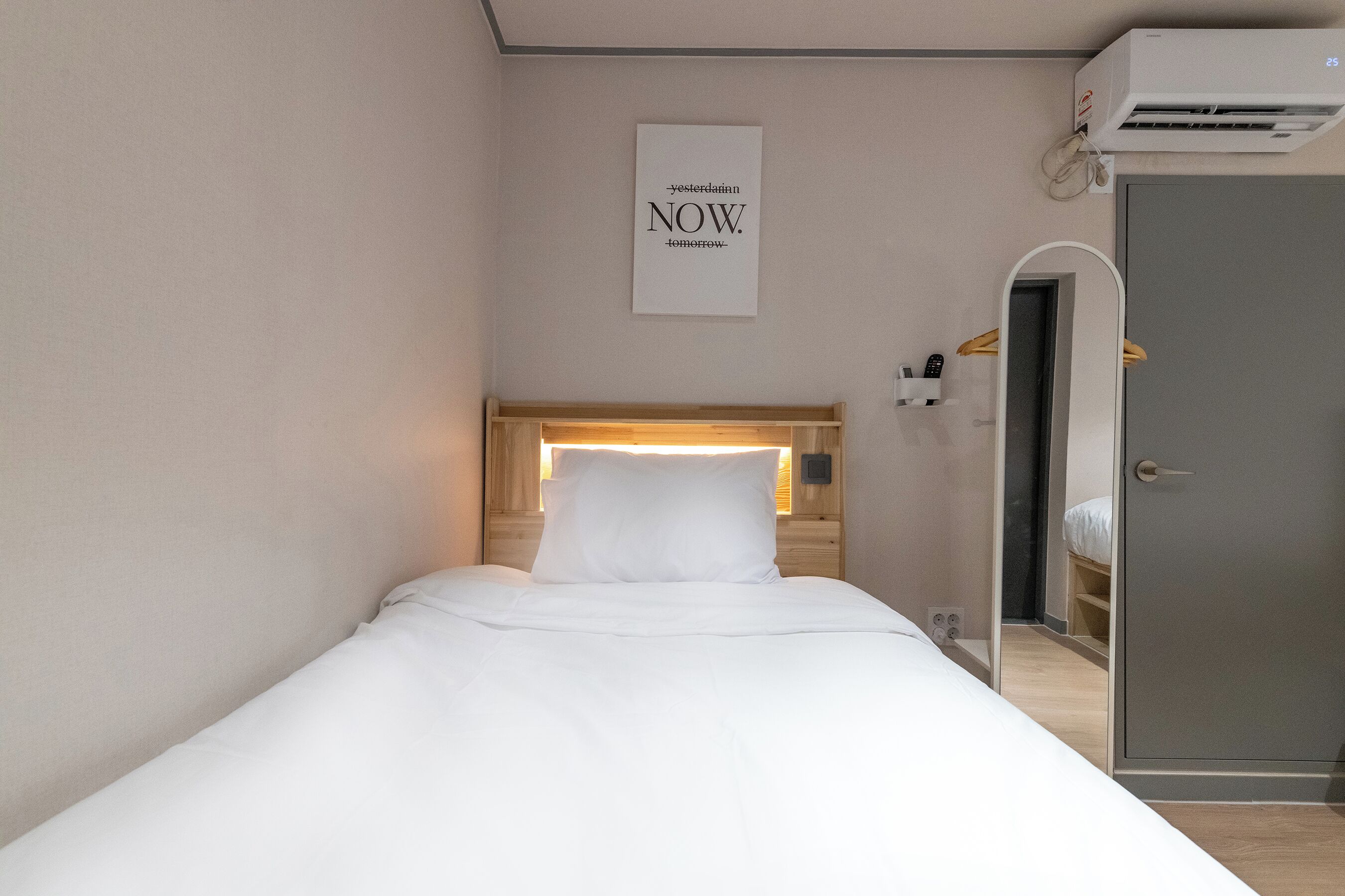 A neatly made bed with a white headboard and pillow, a wall-mounted air conditioner, and a framed sign with text.