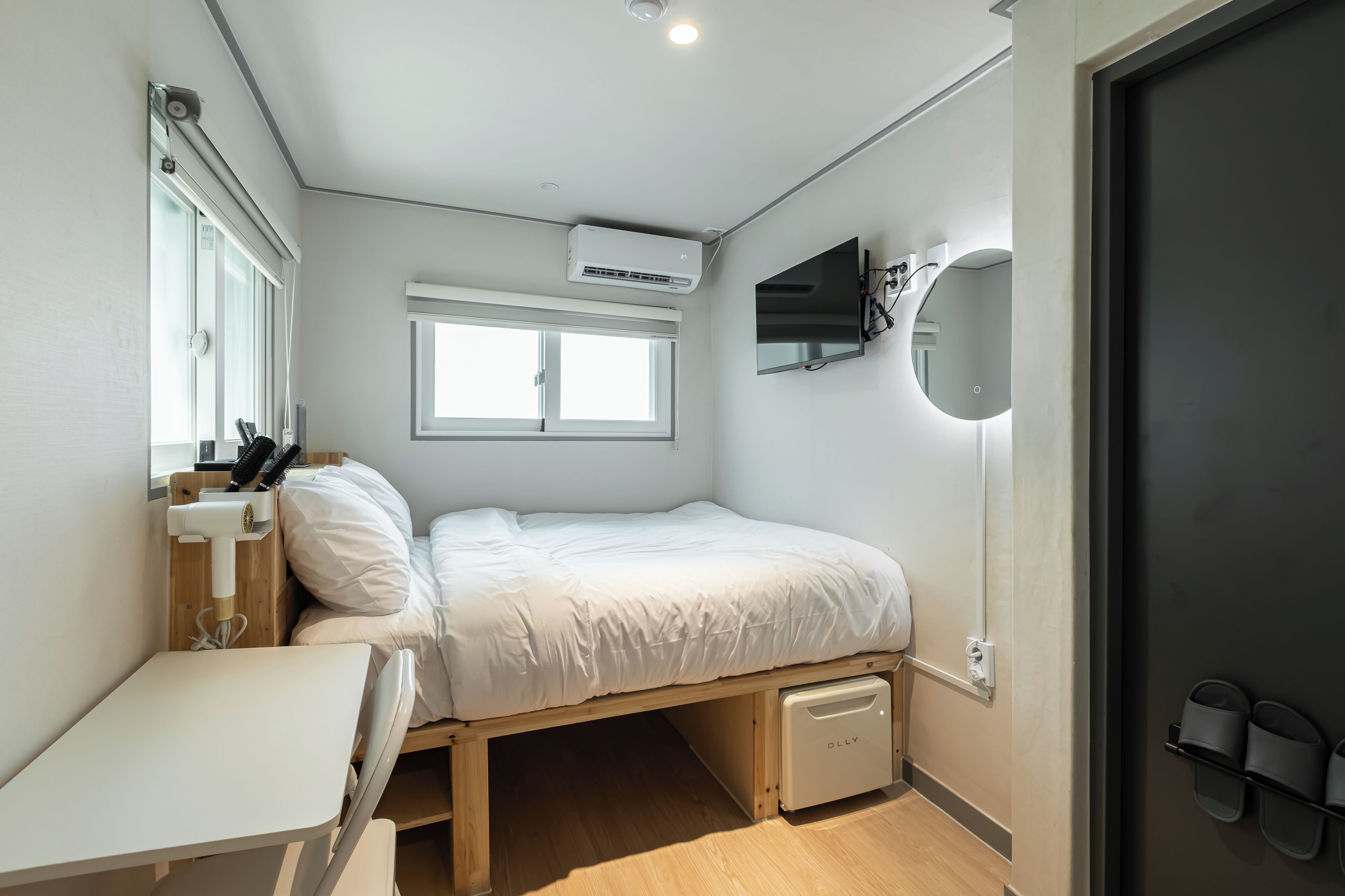 A compact bedroom with a single bed, a small desk, a mirror, and a wall-mounted TV.