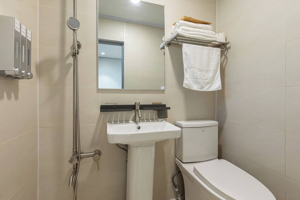 Un baño con lavamanos blanco, inodoro, ducha con cabezal de ducha manual, espejo y toallero con toallas dobladas.