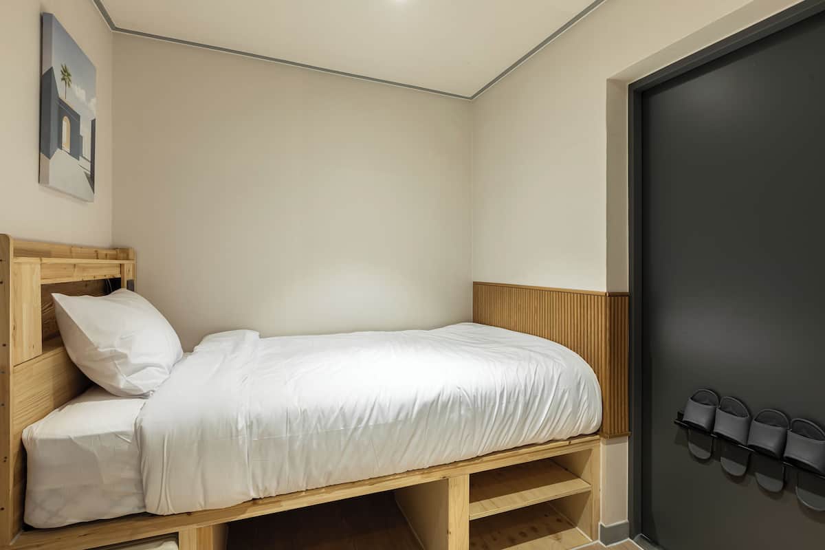 Una cama individual con cabecera de madera, ropa de cama blanca y un zapatero pegado a la pared.