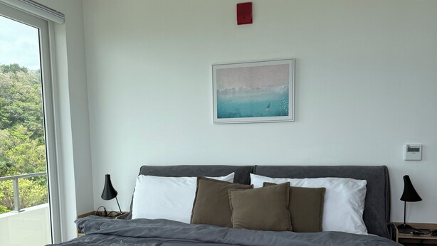Ein Schlafzimmer mit einem großen Bett, einem Nachttisch, einer an der Wand montierten Weckeruhr und einem Fenster mit Blick auf Grünflächen.