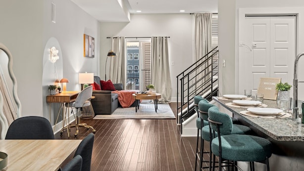 A modern living room with a wooden floor, a sofa, a dining table with chairs, and a kitchen area with a bar counter.