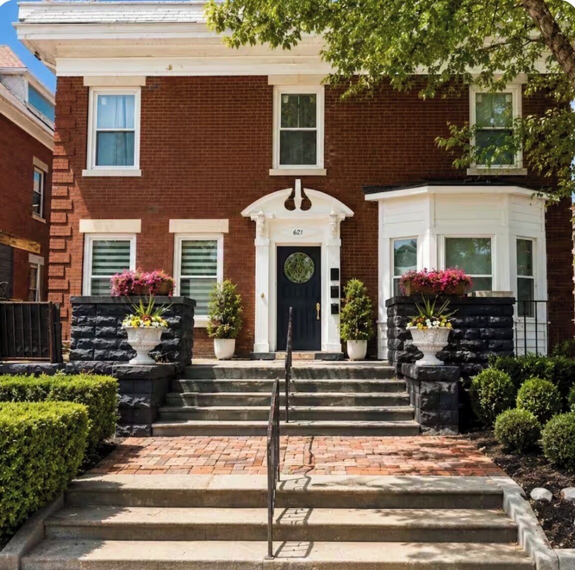 A two-story brick building with a black front door, white trim, and a number '621' above the door. There are two potted plants on either side of the door, and a small garden area with trimmed bushes.