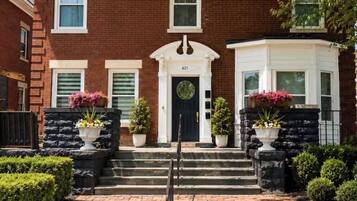 A two-story brick building with a black front door, white trim, and a number '621' above the door. There are two potted plants on either side of the door, and a small garden area with trimmed bushes.