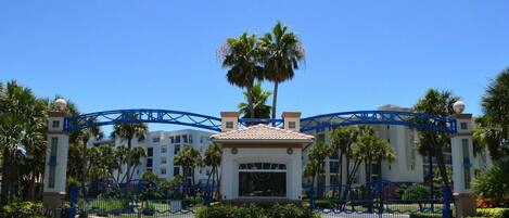 A gated entrance with a clear blue sky, palm trees, and a building with a red-tiled roof.