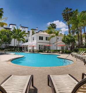 A swimming pool with lounge chairs and umbrellas, surrounded by palm trees and a building with a tiled roof.