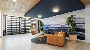 A spacious lobby with a high ceiling, a large mural of a seascape, a wooden floor, a seating area with a sofa and chairs, and potted plants.